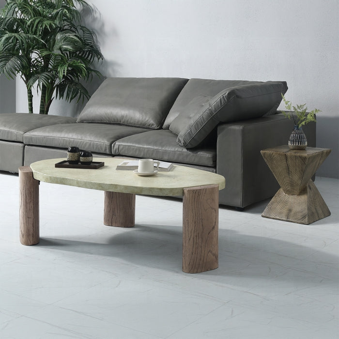white background coffee table three legs cement appearance top with rock shape legs give a wooden earthy appearance  staged in a living room setting with a couch in the background grey and plants 