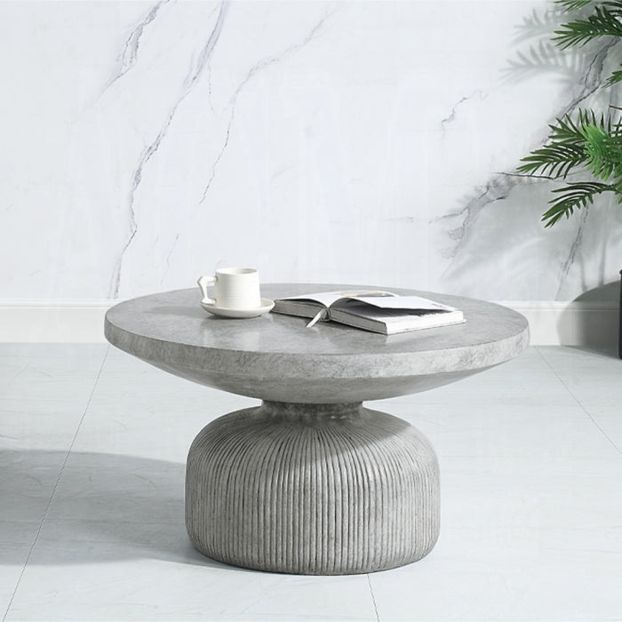 hour glass shape concrete appearance coffee table with ridged lines at the bottom and a round flat top staged with a mug and book ontop 