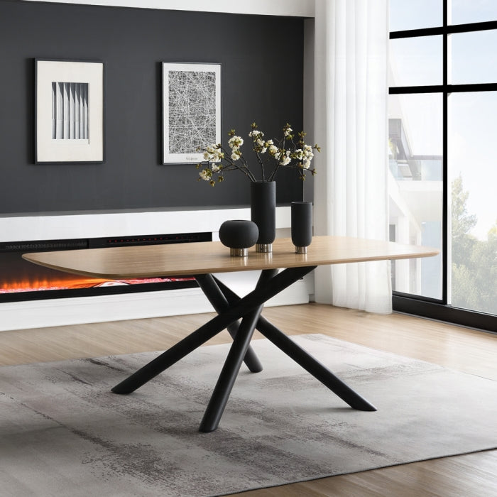 white background light wood color appearance table top rectantgular shape with black criss crossed legs 4 going in different directions staged in a room with flowers on top black vase decor fireplace in the background and a rug underneath the table in light grey 