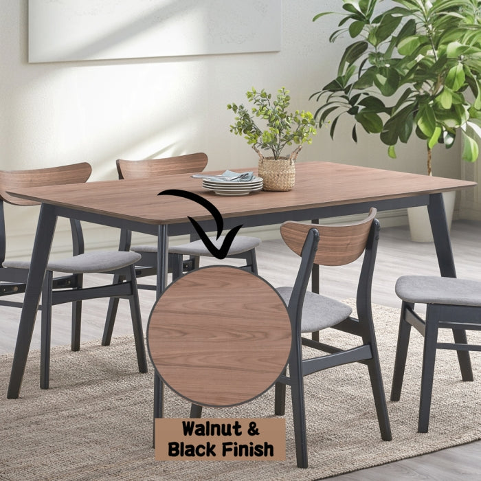 white background dinning table with 4 black legs base and a walnut colored finished as the table top surface staged in a dinning room setting with 4 matching chaise a few plants in the background and a carpet on the floor trying to match color tones with the light greys 