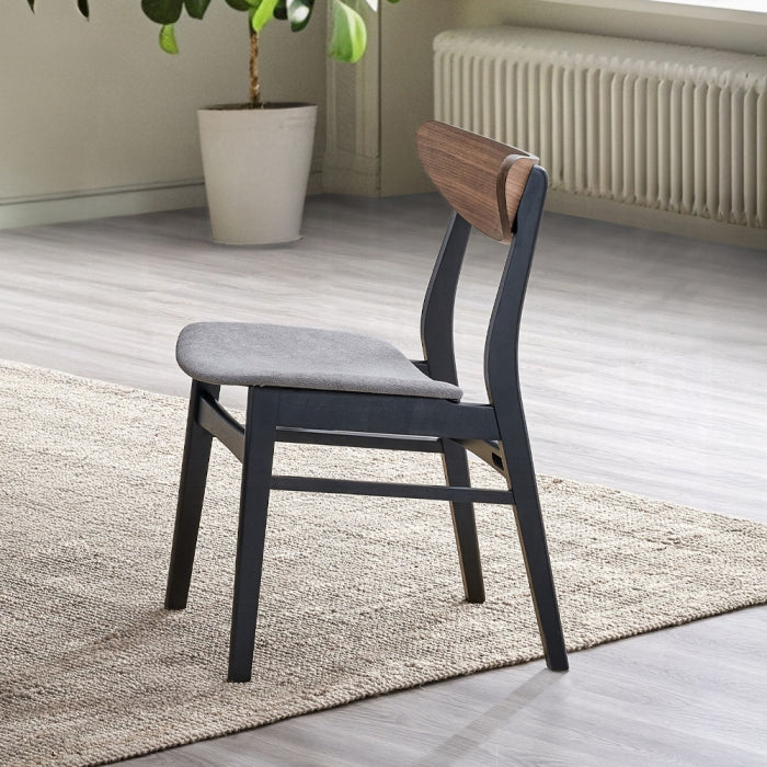 grey padded eat black chair black 4 legs with a wood like medium brown finish as the backrest that is a little curved at the edges staged in a home with grey floors light rug and a plant in the background to show example